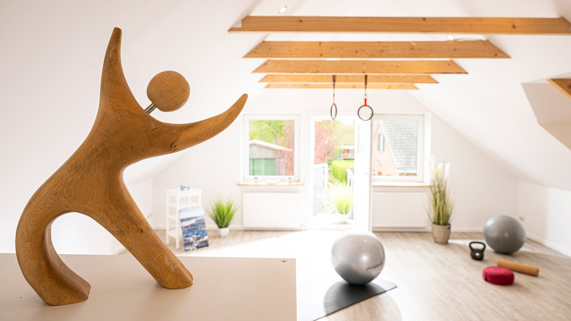 Ein Blick in ein lichtdurchflutetes Behandlungszimmer unter dem Dach. An hölzernen Deckenbalken hängen zwei Turnringe. Im Vordergrund steht eine tanzende Holzfigur mit organischen Formen. Auf dem Holzfußboden liegen Gymnastikbälle und weiteres Trainingsmaterial.